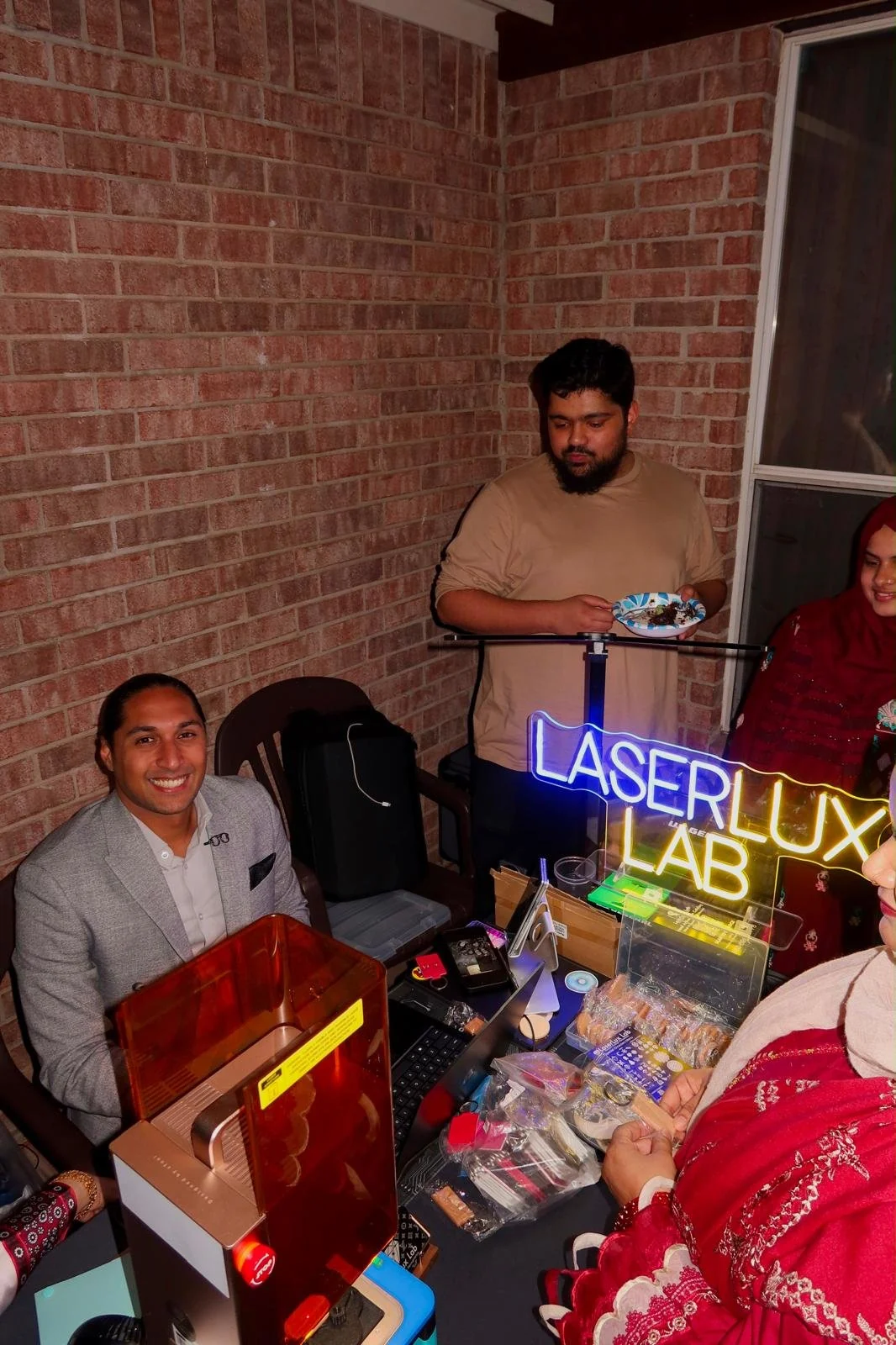The LaserLux Lab founder smiling at a live laser engraving booth with a neon LaserLux Lab sign while guests look on